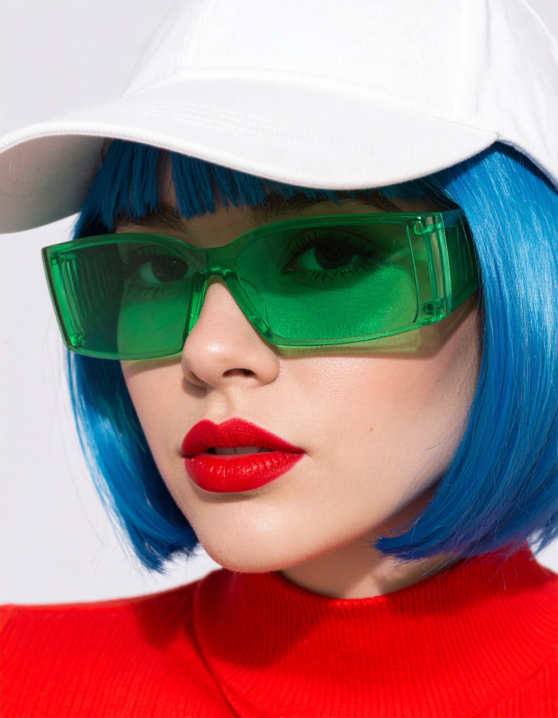 Close-up of a fashion model with blue hair, wearing neon green translucent visor-style sunglasses, a white cap, and a red high-neck minimalistic dress. Shot in bright midday sunlight with strong shado